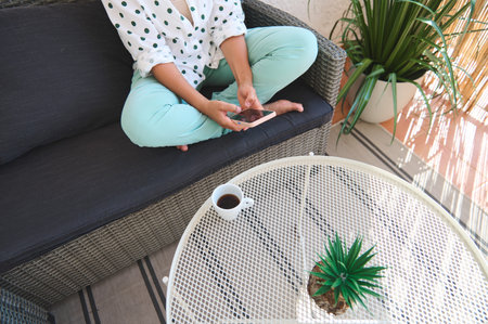 A woman sitting cross-legged on a patio sofa using her smartphone, with a cup of coffee and a plant...
