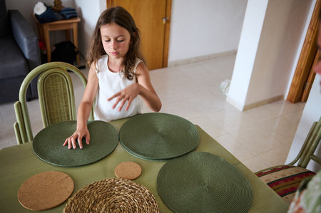 A young girl in a white dress arranges green placemats on a dining table in a cozy home, showcasing concentration and domestic activity.の写真素材