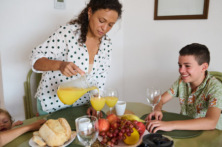 A mother pours fresh orange juice for her children during breakfast. The table is set with various fruits and bread, capturing a warm family morning.の写真素材
