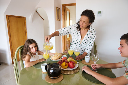 A mother serves orange juice to her children at a breakfast table with fruits and bread, creating a warm family atmosphere.の写真素材