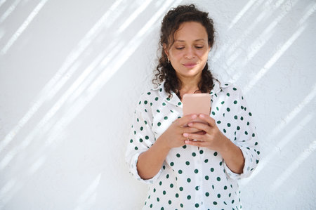 A relaxed woman in a polka dot shirt enjoys using her smartphone, bathed in gentle sunlight against a white background, conveying a sense of calm and connection.の写真素材