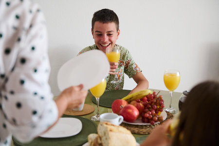 A cheerful morning scene with a young boy enjoying breakfast, surrounded by fresh juice, bread, and fruits, creating a warm family atmosphere.の写真素材