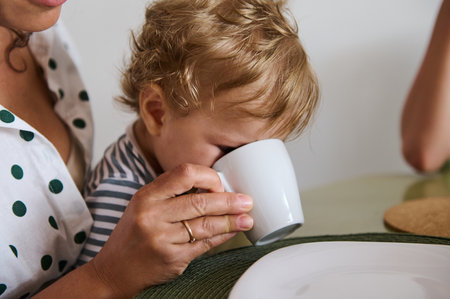 Adorable toddler enjoying a warm drink from a white mug on a parent's lap. Cozy family morning routine.の写真素材