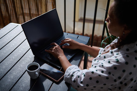A woman types on a laptop while enjoying coffee on a patio. Ideal for remote work, technology, freelance and lifestyle concepts.の写真素材