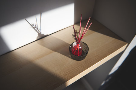 Minimalistic reed diffuser with red sticks on wooden shelf, casting a soft shadow. Evokes warmth, tranquility, and simplicity in home decor.の写真素材