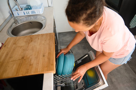 A woman in casual attire loads blue dishes into a dishwasher in a bright kitchen, showcasing the routine of household chores and modern convenience.の写真素材
