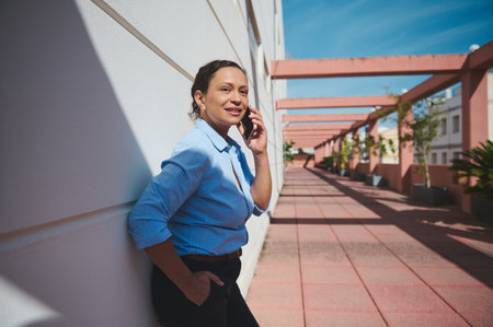Professional woman in a blue shirt having a phone conversation outdoors. She exudes confidence and focus, standing against a sunny architectural backdrop.の写真素材