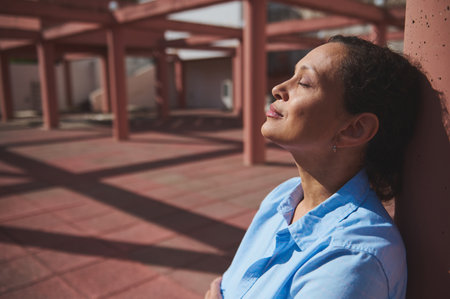 A serene woman in a blue shirt leans against a structure, basking in sunlight with closed eyes, symbolizing relaxation and mindfulness in an urban environment.の写真素材