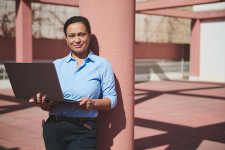 Confident professional woman holding a laptop, standing against a column in bright sunlight, representing success in a contemporary business environment.の写真素材