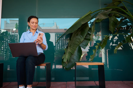 Woman sitting outside using a smartphone and laptop, enjoying a relaxing moment in a modern outdoor space with lush plants.の写真素材