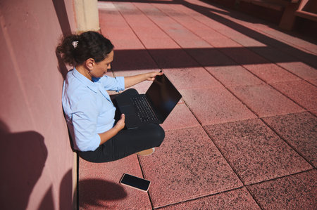 A woman in business casual attire works on a laptop outdoors, sitting in the shade. The scene conveys remote work and digital nomad flexibility.の写真素材