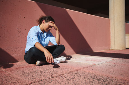 A young woman sits alone against a pink wall, showing signs of stress and contemplation in a sunny urban setting, casting shadows on the ground.の写真素材