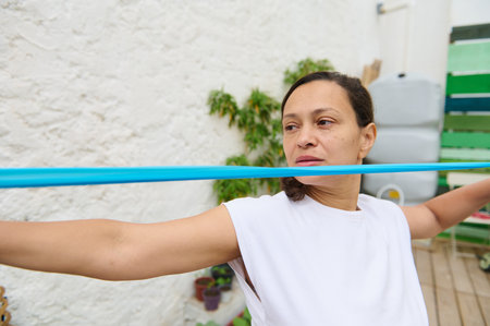 A woman in a white shirt uses an exercise band for stretching in an outdoor garden, emphasizing...