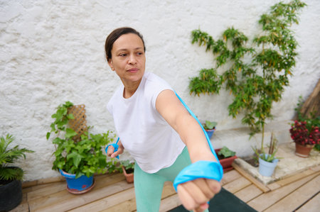 Focused woman using a resistance band for exercise in a garden setting. Embracing a healthy...