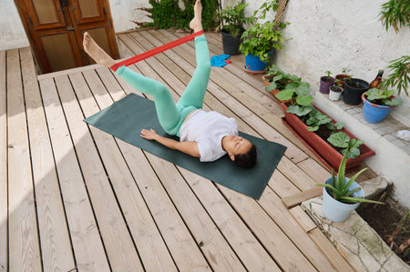 A woman performs yoga using a resistance band on a sunlit wooden deck, enhancing fitness and...