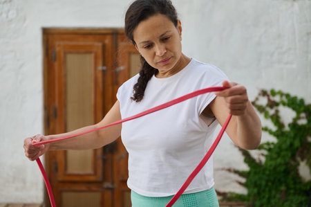 A woman in a white t-shirt uses a resistance band for stretching exercises outdoors, promoting...