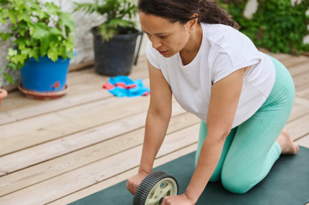 A woman exercises with an ab roller on a wooden deck, emphasizing fitness and strength in an outdoor setting adorned with potted plants.の写真素材