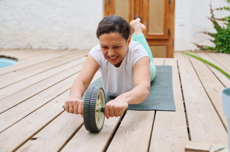 A woman on a yoga mat doing ab rollouts with a wheel outdoors, emphasizing fitness, core strength, and determination in a natural setting.の写真素材