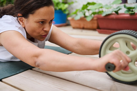 A determined woman exercises with an ab roller on a deck, blending fitness with nature in an outdoor setting filled with plants.の写真素材