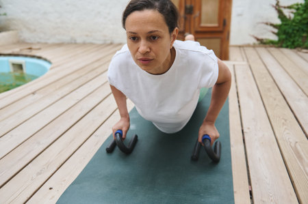 A determined woman performing push-ups using push-up bars outdoors, emphasizing strength and fitness...