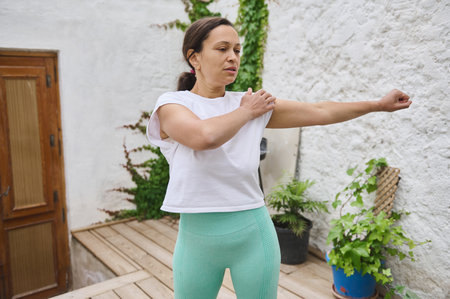 A woman performs an arm stretch in a tranquil outdoor setting, wearing sporty attire, promoting a...