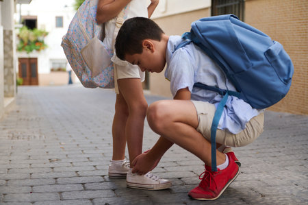 A young boy kneels to help his friend tie her shoelaces on a cobblestone street. Both wear backpacks, displaying friendship and cooperation on the way to school.の写真素材