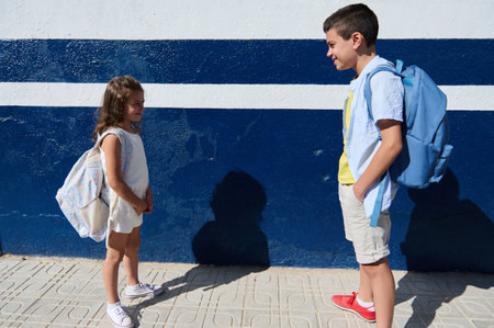 A young girl and a boy stand in sunlight, wearing backpacks and casual outfits, sharing a friendly...
