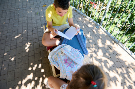 Two children sharing a book while sitting on a park bench. The scene captures a quiet moment of focus and learning outdoors on a sunny day with backpacks nearby.の写真素材