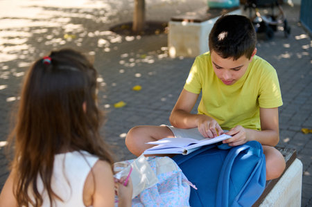 Two kids sitting on a bench, engaged in studying from a book, outdoors. The scene conveys learning and friendship.の写真素材