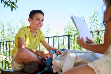 A smiling boy in a yellow shirt sits outdoors, engaging in a casual study session with a friend...