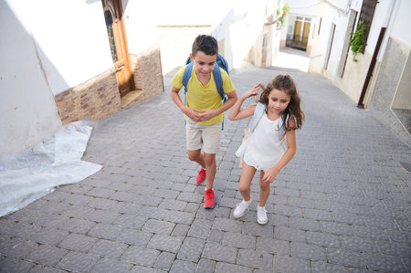 A joyful brother and sister duo walk up a charming street on a sunny day, wearing casual clothes and backpacks, enjoying their outdoor adventure.の写真素材
