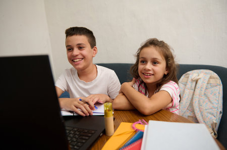 Two cheerful kids engaged in online education, smiling at a laptop screen, with notebooks and school supplies nearby.の写真素材