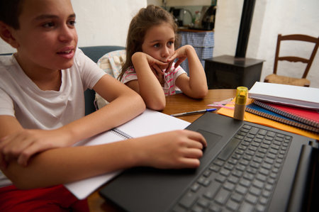 Two children engage in home learning, using a laptop and notebooks. They focus on educational tasks...