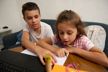 Young siblings engaged in learning at home, working together on schoolwork with a laptop and notebook on a table.の写真素材