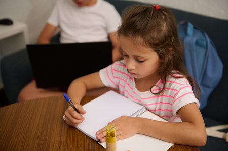 A young girl focused on studying and writing in a notebook, with a boy working on a laptop in the background.の写真素材
