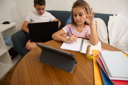 Two children are participating in remote learning activities. One uses a tablet, the other a laptop,...