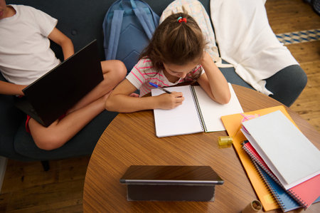 Two children engaged in learning at home, one writing in a notebook and the other using a laptop, creating a focused study environment.の写真素材