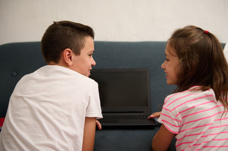 Two smiling siblings enjoy time together on the couch, bonding and sharing fun moments while using a laptop.の写真素材