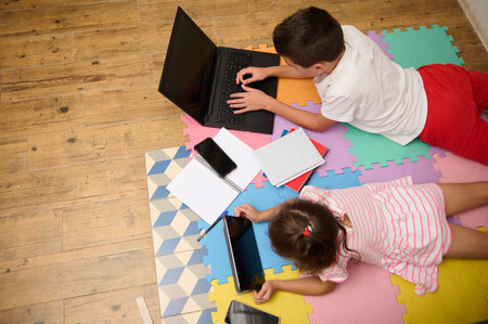 Two children using a laptop and tablet for online learning while lying on colorful foam mats. Nearby, notebooks and pens indicate a study session.の写真素材