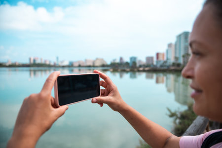 A woman takes a photo of a city skyline with her smartphone, showcasing serene urban waterside scenery. The image conveys travel, technology, and exploration.の写真素材