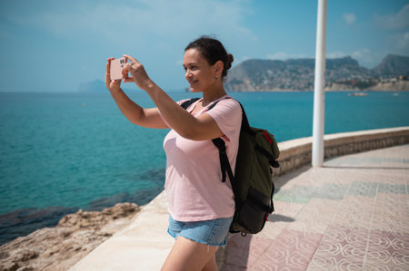 A woman smiles while taking a selfie on a sunny coastal walkway, capturing the scenic view. She's wearing a backpack and casual attire, enjoying a day at the seaside.の写真素材