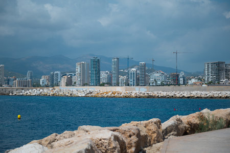 A panoramic view of a coastal city featuring modern skyscrapers, calm blue waters, and mountains in the background, under a cloudy sky.の写真素材