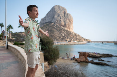 A happy boy with open arms enjoys a beautiful sunny day near a scenic coastline with a rocky mountain backdrop.の写真素材