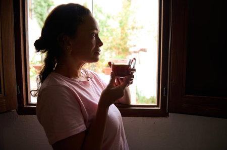 Woman holding a coffee cup, gazing out a window with a thoughtful expression. The warm light creates a peaceful atmosphere.の写真素材