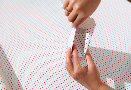 Hands wrapping a small polka dot gift box on white paper under bright lighting, showcasing holiday preparation.の写真素材