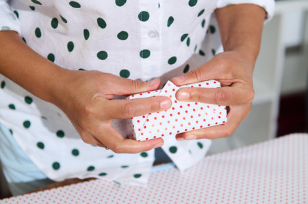 Close-up of hands wrapping a small gift in red polka dot paper, focusing on the art of present...