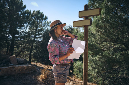 A woman in a hat examines a map and signpost while hiking in a forest, contemplating her path through the scenic landscape.の写真素材