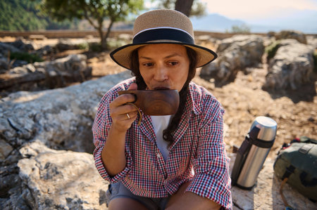 A woman enjoys a hot drink during a peaceful outdoor hike, wearing a straw hat and plaid shirt,...