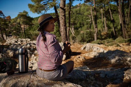 Woman enjoying peaceful forest scene with a cup of tea, seated on rocky terrain. Sunlight filters through trees, creating a calm outdoor setting.の写真素材
