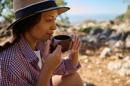 Woman in a checkered shirt and hat enjoying a cup of coffee in a serene nature setting. Relaxation and tranquility in the great outdoors.の写真素材